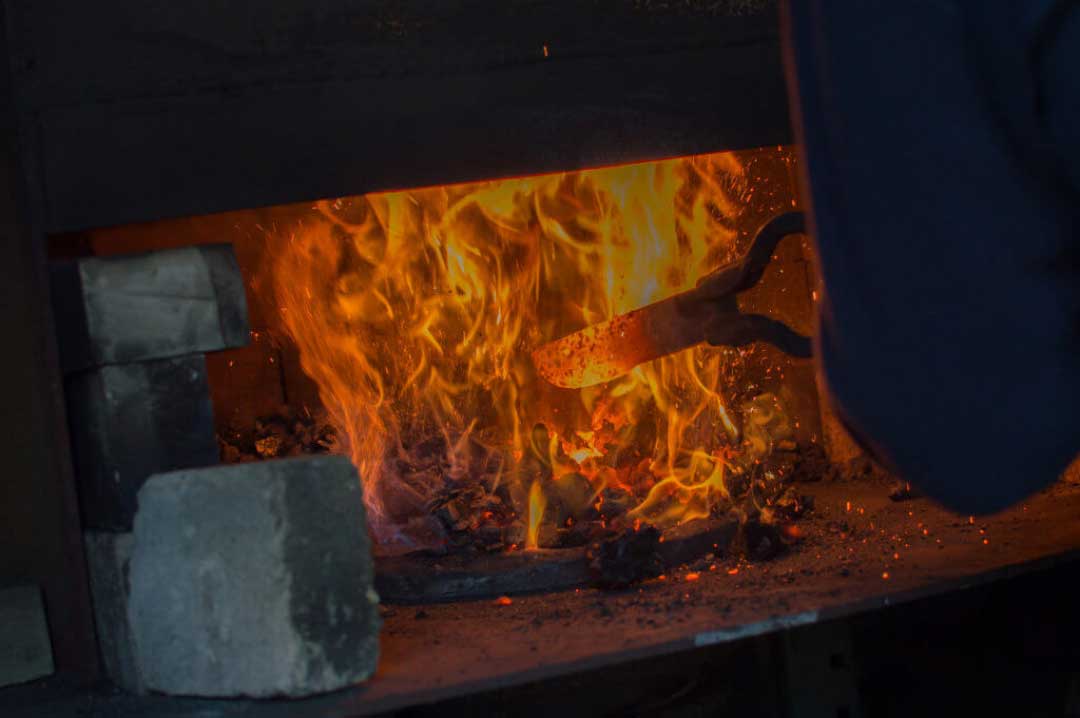 Person using tongs to handle hot metal in a forge with flames and sparks.