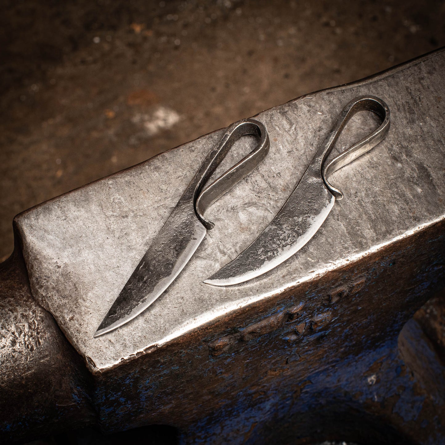 Two handcrafted knives with engraved details, resting on a metallic surface, likely an anvil, with a blurred background.